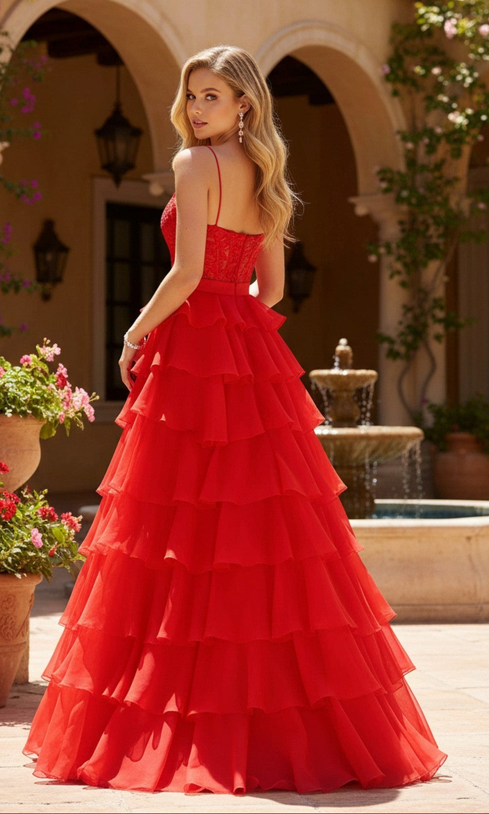 Wearing the PromGirl Exclusive 250202 long prom dress, a woman in red stands outdoors near a fountain, her back to the camera as she turns her head to the side.